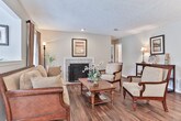 This single-story home in Riverdale, Georgia offers a well-organized layout with distinct living spaces connected by consistent design elements throughout. Dark hardwood-style flooring flows across the main living areas, creating a cohesive and grounded aesthetic that complements the neutral gray wall tones and white trim.

The living room serves as a central gathering space, anchored by a tile-surround fireplace and furnished with warm, earthy tones. Natural light enters through windows dressed with linen-style drapes and horizontal blinds. A half-wall partition separates the living room from adjacent areas, adding architectural interest while maintaining an open feel.

The dining area is a defined yet intimate space, accessible from the living room and positioned near the kitchen. It features the same hardwood-style flooring and overhead lighting, making it well-suited for everyday dining.

The kitchen is equipped with dark wood cabinetry, white countertops, a double-basin sink, a white range, and a dishwasher, offering practical functionality. Under-cabinet lighting provides focused illumination over the work surfaces.

The bedroom features plush carpeting and a textured ceiling, providing a quieter, softer retreat. The full bathroom includes a single-vanity with dark wood cabinetry, a tub-shower combination, and hardwood-style flooring. A utility area houses the water heater and electrical panel, enclosed behind louvered doors.
