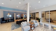 Located at 1660 Metro in Chula Vista, this single-floor business center offers a thoughtfully designed professional environment that blends functionality with contemporary style. The space is anchored by a spacious main lounge featuring light tile flooring, recessed lighting, and large floor-to-ceiling windows that flood the area with natural light and provide views of the surrounding exterior. Comfortable seating arrangements — including plush sofas, armchairs, and patterned area rugs — create a welcoming atmosphere for casual meetings or relaxation. A statement chandelier with exposed globe bulbs adds a modern design touch overhead. The workspace transitions into a dedicated co-working area with a long countertop bar lined with sleek black chairs, bathed in natural light from a full window wall, and accented by bold sunburst wall décor on a slate-blue feature wall. Booth-style seating with individual monitor screens offers semi-private work alcoves. A fully enclosed glass conference room provides a professional setting equipped with a central meeting table, wall-mounted display, and whiteboard. The entry corridor features a geometric accent wall and polished elevator access, completing the center's clean, modern aesthetic.