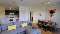 This single-floor space offers a thoughtfully designed open-concept layout that seamlessly connects the living, dining, and kitchen areas into one cohesive and functional environment. The living area is anchored by a spacious sectional sofa in cool gray tones, complemented by yellow accent pillows that add a pop of color. A sleek coffee table centers the seating area, and decorative circular mirrors arranged in a cluster adorn the white walls, adding visual interest and depth. Track lighting runs along the ceiling, providing adjustable, well-distributed illumination throughout the space. The dining area sits adjacent to the living room and features a warm-toned oval dining table surrounded by modern chairs, creating an inviting space for shared meals. The kitchen is partially open to the main living area and is outfitted with dark cabinetry, stainless steel appliances, and a granite countertop breakfast bar, offering both style and practicality. Rich hardwood flooring flows throughout the main areas, adding warmth and continuity to the overall aesthetic. The clean white walls and neutral palette allow the furnishings and accent pieces to stand out, while the open layout ensures a bright, airy feel. A hallway leading to additional rooms is visible in the background, suggesting further private spaces beyond the main living area.