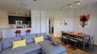 This single-floor space offers a thoughtfully designed open-concept layout that seamlessly connects the living, dining, and kitchen areas into one cohesive and functional environment. The living area is anchored by a spacious sectional sofa in cool gray tones, complemented by yellow accent pillows that add a pop of color. A sleek coffee table centers the seating area, and decorative circular mirrors arranged in a cluster adorn the white walls, adding visual interest and depth. Track lighting runs along the ceiling, providing adjustable, well-distributed illumination throughout the space. The dining area sits adjacent to the living room and features a warm-toned oval dining table surrounded by modern chairs, creating an inviting space for shared meals. The kitchen is partially open to the main living area and is outfitted with dark cabinetry, stainless steel appliances, and a granite countertop breakfast bar, offering both style and practicality. Rich hardwood flooring flows throughout the main areas, adding warmth and continuity to the overall aesthetic. The clean white walls and neutral palette allow the furnishings and accent pieces to stand out, while the open layout ensures a bright, airy feel. A hallway leading to additional rooms is visible in the background, suggesting further private spaces beyond the main living area.