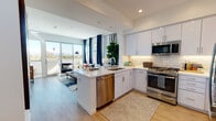 This thoughtfully designed single-floor residence combines modern aesthetics with functional living. The open-concept layout seamlessly connects the kitchen and living area, creating a bright and inviting atmosphere. The kitchen is outfitted with crisp white cabinetry, quartz countertops, a classic subway tile backsplash, and a full suite of stainless steel appliances, including a gas range, built-in microwave, dishwasher, and refrigerator. A peninsula with bar seating offers additional dining and prep space. The living area is bathed in natural light from floor-to-ceiling sliding glass doors that open to a private balcony, framing open city views. Navy blue curtains add a bold, stylish accent against the neutral palette. Light wood-tone flooring flows consistently throughout the space. The bedroom is a comfortable retreat, featuring a ceiling fan, a generous window with city views, and ample room for furnishings. The bathroom is clean and contemporary, with a white vanity, quartz countertop, illuminated mirror, built-in shelving, and a shower-tub combination with white tile surround. The entryway provides a welcoming transition into the home, accented by a transom window above the front door that draws in additional natural light.