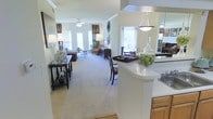 This single-floor residence showcases a thoughtfully designed open-concept layout that seamlessly connects the kitchen and living areas. The kitchen features warm wood cabinetry, a sleek white countertop peninsula with a decorative pendant light overhead, and a mirrored backsplash that adds depth and brightness to the space. A built-in cooktop and generous counter space make it both functional and stylish.

The open layout flows effortlessly into a generously sized living area finished with plush wall-to-wall carpeting that adds warmth underfoot. Crown molding lines the upper walls, lending a refined architectural touch throughout. The living room is bathed in natural light streaming through multiple large windows dressed with elegant curtain panels, creating a bright and welcoming ambiance.

A ceiling fan centered in the living area ensures comfort year-round, while a decorative console table along the entryway wall provides a graceful transition into the main space. The room is furnished with classic, coordinated pieces including upholstered seating and accent tables, conveying a comfortable and polished atmosphere. The overall design balances practicality with style, making this a versatile and inviting single-level home.