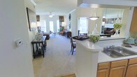 This single-floor residence showcases a thoughtfully designed open-concept layout that seamlessly connects the kitchen and living areas. The kitchen features warm wood cabinetry, a sleek white countertop peninsula with a decorative pendant light overhead, and a mirrored backsplash that adds depth and brightness to the space. A built-in cooktop and generous counter space make it both functional and stylish.

The open layout flows effortlessly into a generously sized living area finished with plush wall-to-wall carpeting that adds warmth underfoot. Crown molding lines the upper walls, lending a refined architectural touch throughout. The living room is bathed in natural light streaming through multiple large windows dressed with elegant curtain panels, creating a bright and welcoming ambiance.

A ceiling fan centered in the living area ensures comfort year-round, while a decorative console table along the entryway wall provides a graceful transition into the main space. The room is furnished with classic, coordinated pieces including upholstered seating and accent tables, conveying a comfortable and polished atmosphere. The overall design balances practicality with style, making this a versatile and inviting single-level home.