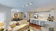 This thoughtfully designed single-floor residence showcases a seamless open-concept layout that connects the living, dining, and kitchen areas into one cohesive and inviting space. Rich hardwood flooring flows throughout, anchoring the warm yet contemporary aesthetic.

The kitchen is a standout feature, boasting crisp white cabinetry, a large central island with a quartz countertop and bar seating, pendant lighting, and stainless steel appliances including a French door refrigerator. A classic subway tile backsplash adds a refined finishing touch.

The living area is generously proportioned, illuminated by a sleek track lighting system mounted on a smooth white ceiling. The space comfortably accommodates a plush sofa ensemble, layered with neutral and gold-toned accents that complement the overall design palette.

Adjacent to the living room, the dining area provides an intimate setting, complete with a round decorative mirror that adds depth and character to the wall.

Throughout the space, neutral gray and white tones create a bright, airy atmosphere, while gold and warm earth accents lend a sense of elegance. Multiple doorways visible from the main living area suggest additional private rooms beyond, offering both functionality and comfortable living in a well-appointed, modern floor plan.