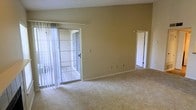 This single-floor residential space offers a clean, functional layout designed for comfortable everyday living. The main living area is carpeted in a neutral, light-toned material that adds warmth underfoot and complements the beige-painted walls. A set of sliding glass doors, fitted with vertical blinds, opens to an outdoor balcony or patio, allowing natural light to filter into the room and providing easy access to the exterior. The textured ceiling adds subtle visual interest overhead, while the simple, open wall space offers flexibility for furnishing and décor. A doorway on the far side of the room connects to an additional area of the space, suggesting a practical, connected floor plan that encourages easy movement throughout. The overall ambiance is neutral and move-in ready, presenting a blank canvas that is well-suited for personalization. The space benefits from an abundance of natural light through the glass door access point, creating an airy and open atmosphere despite its modest scale. Clean lines, minimal architectural detailing, and a cohesive neutral color palette define the character of this straightforward, well-maintained residential space.