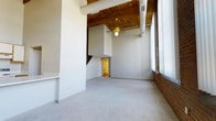 This two-story loft-style residence offers a compelling mix of industrial heritage and contemporary living across two well-appointed levels. The main living area immediately impresses with its soaring ceilings, where rich wood beams and planking provide warm contrast against crisp white walls. Exposed cylindrical ductwork reinforces the industrial aesthetic, lending an authentic loft character to the space.

Large windows along the brick-accented wall flood the living area with natural light, highlighting the neutral carpet flooring and open layout below. The original brick wall adds texture and a sense of history, anchoring the space with architectural depth. A staircase, partially visible from the main room, connects the two floors, with a glimpsed lower-level bathroom illuminated in warm tones suggesting thoughtful interior lighting throughout.

The two-bedroom, two-bathroom layout provides practical functionality, with the dual-floor arrangement offering a natural separation of living and sleeping areas. The overall ambiance balances raw, urban character with a livable warmth, making it well-suited for those who appreciate architecturally distinctive spaces with a nod to industrial design heritage.