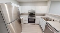 This 640-square-foot, single-floor, one-bedroom, one-bathroom unit offers a well-organized layout designed for comfortable living. The combined kitchen and living area forms the heart of the home, featuring light wood-look flooring, recessed lighting, and multiple windows with blinds that allow ample natural light. The kitchen is equipped with stainless steel appliances — including a refrigerator, range, and dishwasher — complemented by white shaker-style cabinetry, light countertops, and a double basin sink. The living area provides direct access to the outdoors via a side door, adding convenience and additional light. The 140-square-foot bedroom features plush carpeting, a natural light window, and ceiling light fixture, with direct access to a dedicated walk-in closet outfitted with wire shelving systems. The bathroom is thoughtfully designed with accessibility in mind, featuring multiple grab bars around the tub/shower combo and toilet area, set against light tile flooring and neutral walls. A dedicated laundry room with washer/dryer hookups and wire shelving above rounds out this practical and efficiently designed unit.