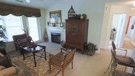 This single-story space showcases a thoughtfully designed living area with vaulted ceilings that lend an open, airy feel to the room. The focal point is a clean white fireplace framed by a traditional mantel, accented with decorative candlesticks and framed artwork above. Natural light filters in through a window dressed with woven bamboo-style valances, complementing the soft, sage-green walls and neutral carpeted flooring.

The room features an eclectic mix of furnishings, including a carved wooden armchair with woven seating, a dark metal side table, and a richly patterned area rug that anchors the space. A substantial mahogany-toned cabinet with brass hardware serves as a prominent storage and display piece, topped with a decorative birdcage and ornamental spheres, adding height and visual interest. Greenery in the form of potted plants and botanical accents throughout the room brings warmth and a natural element to the overall aesthetic. The combination of textures — woven, wood, and metal — creates a layered, globally inspired ambiance that feels both inviting and curated.