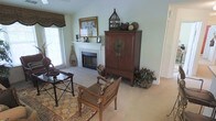 This single-story space showcases a thoughtfully designed living area with vaulted ceilings that lend an open, airy feel to the room. The focal point is a clean white fireplace framed by a traditional mantel, accented with decorative candlesticks and framed artwork above. Natural light filters in through a window dressed with woven bamboo-style valances, complementing the soft, sage-green walls and neutral carpeted flooring.

The room features an eclectic mix of furnishings, including a carved wooden armchair with woven seating, a dark metal side table, and a richly patterned area rug that anchors the space. A substantial mahogany-toned cabinet with brass hardware serves as a prominent storage and display piece, topped with a decorative birdcage and ornamental spheres, adding height and visual interest. Greenery in the form of potted plants and botanical accents throughout the room brings warmth and a natural element to the overall aesthetic. The combination of textures — woven, wood, and metal — creates a layered, globally inspired ambiance that feels both inviting and curated.