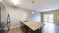 This efficiently designed 1-bedroom, 1-bathroom unit offers a thoughtful layout across a single floor. The open-concept kitchen and living area serves as the heart of the unit, where a central island with a stone-look countertop and double stainless steel sink acts as a natural divider between the two spaces. Pendant lighting above the island adds a modern touch, while dark espresso cabinetry and black appliances — including a refrigerator, range, and built-in microwave — provide a sleek, cohesive aesthetic. Durable wood-look flooring runs throughout the main living areas, complemented by warm neutral-toned walls and baseboard heating.

The bedroom is a comfortable, carpeted retreat featuring a window that brings in natural light and a view of the surrounding neighborhood. Its understated design offers a calm and flexible space.

The full bathroom is finished with a granite-top vanity, dark cabinetry matching the kitchen, a full tub-shower combination with a curved curtain rod, and neutral tile flooring. A dedicated laundry room is equipped with washer and dryer hookups, a wire storage shelf above, and tile flooring, adding practical everyday convenience to this compact yet functional home.