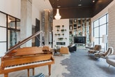 Located in Town Square, Alpharetta, GA, this single-floor communal space presents a refined blend of contemporary and transitional design. Soaring ceilings anchored by exposed concrete columns define the grand scale of the main lounge area, which is illuminated by elegant crystal chandeliers that contrast beautifully with the industrial architectural framework. The flooring transitions seamlessly from light hardwood to large-format hexagonal tile, delineating distinct zones throughout the space. A focal wall features a fireplace flanked by a floor-to-ceiling built-in bookshelf filled with curated books and trailing greenery, alongside a mounted television, creating an inviting library-lounge atmosphere. Seating arrangements include plush white sofas, leather accent chairs, and bold houndstooth-upholstered armchairs positioned near oversized windows that flood the space with natural light and offer views of the surrounding Town Square streetscape. A glass-walled conference room with steel-framed partitions provides a transparent, professional meeting environment visible from the main lounge. Additional amenities include a coffee bar area and a grand piano, lending the space a cultured, welcoming character suited for both social gatherings and productive work.