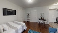 Located at 2220 Walnut Street in Philadelphia, PA, this single-floor studio unit offers a well-organized layout that maximizes every inch of available space. The main living area features rich, dark hardwood flooring that anchors the room, complemented by neutral white walls and a textured ceiling. A blue sofa and coordinating area rug define the living zone, which flows seamlessly into the sleeping area — demonstrating the efficient, open-plan design of the unit. Two interior doors in the bedroom area provide closet access, while a wall-mounted television and compact shelving unit add to the room's functionality. Natural light enters through a window fitted with white blinds, brightening the space throughout the day. The kitchen is tucked neatly off the main area and is outfitted with dark espresso cabinetry, dark granite countertops, a stainless steel gas range, an over-range exhaust hood, a stainless steel sink, and a full-size stainless steel refrigerator — all set against a light tile backsplash. The bathroom features a clean, bright design with a bathtub-shower combination adorned with white subway tile and decorative mosaic border accents in black and neutral tones, adding a refined touch to the utilitarian space.