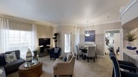 This single-floor residence offers a thoughtfully designed open-concept layout that seamlessly connects the living and dining areas, creating an inviting and functional living environment. The living area is generously sized, featuring plush wall-to-wall carpeting, crisp white crown molding, and neutral-toned walls that lend a clean, timeless aesthetic. Large windows dressed with white curtains and blinds allow abundant natural light to fill the space, enhancing its warm and airy ambiance. A wall-mounted flat-screen television anchors the living area, which is complemented by comfortable seating and tasteful décor, including a tall indoor plant that adds a natural, refreshing touch. The dining area flows naturally from the living space and is centered around a round dining table paired with upholstered chairs, positioned beneath an elegant pendant light fixture. A framed abstract artwork adorns the wall above the dining area, adding a stylish focal point. A glass-paneled door provides access to the exterior, further brightening the space. The overall design strikes a balance between comfort and sophistication, making it a welcoming and practical home.