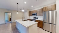 This single-floor unit showcases a contemporary design with a neutral color palette and light hardwood-style flooring throughout the main living areas. The open-concept layout connects the living room to the kitchen, which serves as the centerpiece of the space. The kitchen is equipped with walnut-toned cabinetry, a white subway tile backsplash, stainless steel appliances including a French door refrigerator, built-in microwave, and electric range. A large central island with a granite countertop and undermount sink is illuminated by stylish pendant lights, offering both functionality and visual appeal. An in-unit washer is accessible just off the kitchen area. The living room features light hardwood-style flooring, a ceiling fan, and sliding glass doors that open to a private balcony. Bedrooms are finished with plush carpet and ceiling fans, with natural light provided by sizable windows. A spacious walk-in closet with built-in shelving and hanging rods offers generous storage. Two bathrooms are present: one features a soaking tub and wood-toned vanity with a granite countertop, while the other offers a glass-enclosed walk-in shower with large-format tile, a framed mirror, and matching cabinetry. The overall ambiance is clean, modern, and move-in ready.