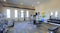 This fitness center offers a practical and well-organized workout environment designed to support a range of exercise routines. The space is finished with multi-toned striped carpeting in blue, green, and yellow hues, adding a dynamic visual element to the room. Recessed lighting in the white ceiling provides consistent illumination throughout, complemented by natural light streaming in through multiple windows along the far wall, creating a bright and inviting atmosphere. A large ventilation duct runs horizontally near the ceiling, reflecting the functional infrastructure of the space. The room is equipped with a cable cross machine, a weight bench, a stationary bike, and a multi-use cable tower, catering to both strength training and cardiovascular workouts. A wall-mounted accessory rack organizes medicine balls, resistance bands, and stability balls in various sizes and colors, keeping the space tidy and accessible. A mounted flat-screen television is positioned for easy viewing during workouts. The neutral wall color and open layout contribute to a clean, focused environment well-suited for daily fitness use.