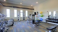 This fitness center offers a practical and well-organized workout environment designed to support a range of exercise routines. The space is finished with multi-toned striped carpeting in blue, green, and yellow hues, adding a dynamic visual element to the room. Recessed lighting in the white ceiling provides consistent illumination throughout, complemented by natural light streaming in through multiple windows along the far wall, creating a bright and inviting atmosphere. A large ventilation duct runs horizontally near the ceiling, reflecting the functional infrastructure of the space. The room is equipped with a cable cross machine, a weight bench, a stationary bike, and a multi-use cable tower, catering to both strength training and cardiovascular workouts. A wall-mounted accessory rack organizes medicine balls, resistance bands, and stability balls in various sizes and colors, keeping the space tidy and accessible. A mounted flat-screen television is positioned for easy viewing during workouts. The neutral wall color and open layout contribute to a clean, focused environment well-suited for daily fitness use.