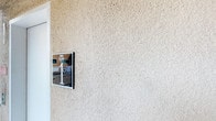 This single-story space is characterized by its textured stucco exterior, finished in a warm, sandy neutral tone that gives it a clean and understated appearance. The rough-cast stucco surface adds depth and visual interest to the facade, a hallmark of southwestern and Mediterranean-influenced architectural styles. Mounted on the exterior wall is a framed sign, suggesting a named property or community entrance point. A glimpse of the surrounding environment through the signage reveals a railing and additional structural elements, hinting at a broader complex or community setting. The overall exterior presents a well-maintained, functional aesthetic consistent with residential or light commercial construction. The single-floor layout contributes to an accessible and straightforward design, with the neutral stucco palette offering a timeless and versatile finish that complements a variety of landscaping and neighboring structures.