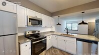 This thoughtfully designed residence spans multiple levels connected by a staircase with a classic blue-painted railing, offering a functional and cohesive layout throughout. The kitchen is a standout feature, outfitted with crisp white shaker-style cabinetry, granite countertops, and a full suite of stainless steel appliances including an electric range, built-in microwave, refrigerator, and dishwasher. An undermount sink with a matte black faucet is positioned at the peninsula, illuminated by stylish dark pendant lights that echo the kitchen's modern hardware accents. Light wood-style luxury vinyl plank flooring runs through the main living areas, lending warmth and continuity. A half bathroom on the main level features a granite-topped vanity with white cabinetry and a framed mirror with refined lighting. Bedrooms are carpeted for comfort, with one room notably offering dual closets for generous storage. A dedicated laundry area houses a full-size washer and dryer tucked behind bifold doors alongside an exterior door access point. Natural light filters through multiple windows fitted with horizontal blinds, keeping the interior bright and welcoming across all rooms.