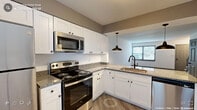 This thoughtfully designed residence spans multiple levels connected by a staircase with a classic blue-painted railing, offering a functional and cohesive layout throughout. The kitchen is a standout feature, outfitted with crisp white shaker-style cabinetry, granite countertops, and a full suite of stainless steel appliances including an electric range, built-in microwave, refrigerator, and dishwasher. An undermount sink with a matte black faucet is positioned at the peninsula, illuminated by stylish dark pendant lights that echo the kitchen's modern hardware accents. Light wood-style luxury vinyl plank flooring runs through the main living areas, lending warmth and continuity. A half bathroom on the main level features a granite-topped vanity with white cabinetry and a framed mirror with refined lighting. Bedrooms are carpeted for comfort, with one room notably offering dual closets for generous storage. A dedicated laundry area houses a full-size washer and dryer tucked behind bifold doors alongside an exterior door access point. Natural light filters through multiple windows fitted with horizontal blinds, keeping the interior bright and welcoming across all rooms.