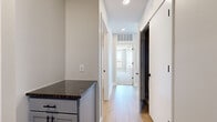 This single-floor residence offers a thoughtfully designed layout across a comfortable footprint, accommodating two bedrooms, two bathrooms, a combined kitchen and living area, a hallway, and two closets. The open-concept kitchen is a standout feature, showcasing grey shaker-style cabinetry, dark granite countertops, stainless steel appliances including a refrigerator, range, and built-in microwave, a peninsula with pendant lighting, and track lighting overhead. Light wood-style flooring runs throughout the main living and kitchen areas, contributing to a clean, contemporary aesthetic. The living area is bathed in natural light through large windows fitted with roller shades, offering views of the surrounding urban landscape. The primary bedroom features soft carpeting and a dedicated closet, while a second bedroom similarly offers carpeted flooring and a window with outdoor views. Both bathrooms are finished with grey vanity cabinetry and wood-look tile flooring; one includes a walk-in shower with chrome fixtures, while the other features a full tub-shower combination. Neutral wall tones and white trim throughout create a cohesive, modern ambiance.