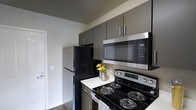 This single-floor space combines a practical layout with contemporary finishes throughout. The open living area is anchored by dark hardwood-style flooring that extends seamlessly through the main spaces, creating a cohesive and polished look. A ceiling fan overhead provides both function and comfort, while a large sliding glass door fitted with vertical blinds allows natural light to fill the room and offers access to the outdoors. An arched doorway with accent lighting provides an elegant transition between living areas, adding a subtle architectural detail to the otherwise clean-lined design.

The kitchen is appointed with dark gray cabinetry featuring sleek modern hardware, providing ample storage space. Stainless steel appliances—including a range, over-the-range microwave, and refrigerator—complement the dark cabinetry and light countertops, creating a sharp contrast that gives the kitchen a contemporary feel. The neutral-toned walls throughout the space contribute to a bright, airy atmosphere. White baseboards and trim offer clean definition between the walls and flooring, tying the overall aesthetic together in a tidy, well-maintained presentation.