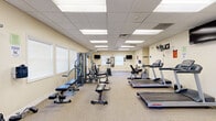 This fitness center at 95 Campus Dr E in Buffalo, New York, offers a comprehensive workout facility within a single, open-plan floor. The space features a drop-panel ceiling with recessed fluorescent lighting that ensures consistent, bright illumination throughout. Warm beige walls and neutral carpet flooring create a clean, welcoming environment conducive to exercise.

The facility is well-stocked with a diverse range of equipment to support both cardiovascular and strength training. Cardio machines include multiple Life Fitness treadmills, a SportsArt elliptical trainer, a stationary upright bike, and a rowing machine, providing ample options for aerobic workouts. For strength training, a multi-station cable machine and adjustable weight benches are available, along with free weight accessories.

Wall-mounted flat-screen televisions are positioned throughout the room, allowing users to stay entertained during their workouts. A wall-mounted clock and full-length mirror add practical functionality to the space. Ceiling fans provide additional air circulation, and large windows fitted with white blinds allow natural light to filter in while maintaining privacy.

The layout affords generous floor space between equipment, ensuring comfortable movement and safe usage. A clearly marked exit door and accessible entry point are also present, contributing to the overall safety and usability of this well-appointed fitness facility.