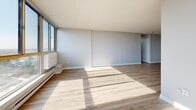 This well-appointed 2-bedroom, 1-bathroom unit offers a thoughtfully designed single-floor layout with a clean, modern aesthetic throughout. Light-toned hardwood flooring runs continuously across the entire space, complementing the bright, neutral color palette of soft whites and cool greys.

The open-concept kitchen and living area forms the heart of the unit. The kitchen is outfitted with sleek, high-gloss white cabinetry, stainless steel appliances including a refrigerator and range, a subway tile backsplash, and a central island with a quartz-style countertop providing additional prep and storage space. The kitchen flows naturally into the living area, which is bathed in abundant natural light streaming through a wide bank of floor-to-ceiling windows offering expansive outdoor views.

Both bedrooms are comfortably sized with light flooring and large windows that bring in generous daylight. The primary bedroom features a wall of sliding panel closets, providing ample storage. One bedroom also benefits from convenient access to an in-unit stacked washer and dryer.

The bathroom is finished in a clean, all-white palette with large-format wall tiles surrounding a full soaking tub and shower, a modern vanity with a stone-look countertop, and tile flooring. A connecting hallway ties all rooms together efficiently, completing this functional and stylish layout.