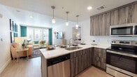 This single-floor residence is designed with a contemporary aesthetic, combining functional layout with modern finishes. The open-concept kitchen serves as a central hub, outfitted with wood-toned cabinetry, quartz countertops, a subway tile backsplash, and a full suite of stainless steel appliances including a dishwasher, range, microwave, and refrigerator. A peninsula with pendant lighting overhead provides bar seating, making it ideal for casual dining or entertaining. Light wood-style flooring flows throughout the main living areas, enhancing the bright, airy atmosphere.

The living room receives generous natural light through large windows dressed with teal curtains, creating a warm and inviting atmosphere. The space comfortably accommodates a full seating arrangement and connects visually to the kitchen for an open feel.

The primary bedroom features large windows, a double-door closet, and carpeted flooring that adds a soft, comfortable feel. The primary bathroom includes a wide vanity with wood-toned cabinetry, quartz countertops, a full-width mirror, and a soaking tub-shower combination. A second bedroom and an additional full bathroom with matching finishes and a tub-shower combo provide comfortable accommodations for guests or additional occupants.