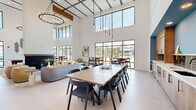 Located at 3036 Philips Highway in Jacksonville, FL, this contemporary community clubhouse delivers an open, airy atmosphere defined by dramatic double-height ceilings and expansive floor-to-ceiling windows. Natural light floods the interior through large black-framed glass panels, offering views of the surrounding palm-lined landscape and connecting the indoor space to its Florida surroundings.

The open-plan layout accommodates multiple functional zones within a single, generously proportioned floor. A central lounge area features a sleek fireplace as its focal point, surrounded by a curated mix of curved sofas, accent chairs in warm tones, and a round cocktail table atop a patterned area rug. Adjacent to this is a large communal dining table with modern black chairs, ideal for group gatherings. Smaller cafe-style tables and counter-height seating near the windows provide additional casual dining options.

A dedicated work and amenity zone includes individual workstations with yellow accent chairs set against a bold teal accent wall, built-in mail and package management stations, and enclosed private office spaces with glass-panel doors. A pool table adds a recreational element to the space.

Overhead, warm wood ceiling beams, an elegant candle-style chandelier, and sleek pendant lighting complement the crisp white walls, creating a sophisticated yet welcoming ambiance throughout.