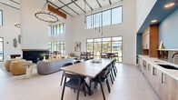 Located at 3036 Philips Highway in Jacksonville, FL, this contemporary community clubhouse delivers an open, airy atmosphere defined by dramatic double-height ceilings and expansive floor-to-ceiling windows. Natural light floods the interior through large black-framed glass panels, offering views of the surrounding palm-lined landscape and connecting the indoor space to its Florida surroundings.

The open-plan layout accommodates multiple functional zones within a single, generously proportioned floor. A central lounge area features a sleek fireplace as its focal point, surrounded by a curated mix of curved sofas, accent chairs in warm tones, and a round cocktail table atop a patterned area rug. Adjacent to this is a large communal dining table with modern black chairs, ideal for group gatherings. Smaller cafe-style tables and counter-height seating near the windows provide additional casual dining options.

A dedicated work and amenity zone includes individual workstations with yellow accent chairs set against a bold teal accent wall, built-in mail and package management stations, and enclosed private office spaces with glass-panel doors. A pool table adds a recreational element to the space.

Overhead, warm wood ceiling beams, an elegant candle-style chandelier, and sleek pendant lighting complement the crisp white walls, creating a sophisticated yet welcoming ambiance throughout.