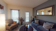 This single-story, two-bedroom, one-bathroom unit offers a practical and comfortable layout across a well-utilized footprint. The living room serves as the central gathering space, featuring carpet flooring, a bold gray accent wall, and multiple windows alongside a glass-paneled door that invites natural light and outdoor views. A ceiling fan is present in the adjoining areas, contributing to the overall airflow throughout the unit.

The entryway provides a welcoming transition into the home, with wall decor and a storage console flanking the front door. The kitchen is efficiently designed in an L-shaped configuration, offering ample white cabinetry, light wood-style flooring, black appliances including a refrigerator and electric range, and a built-in breakfast bar that opens toward the living area.

Both bedrooms are carpeted and receive natural light through large windows with blinds. The primary bedroom features a gray accent wall, ceiling fan, and generous floor space, while the second bedroom includes a closet and flexible layout potential. The bathroom offers a full tub-shower combination with white tile surround and hardwood-style flooring, accompanied by a vanity with mirror and ample counter space.