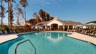 Located at 1090 Summersweet Dr in New Bern, North Carolina, this single-story outdoor amenity complex centers around a striking freeform swimming pool with a distinctive figure-eight shape and crystal-clear blue water. The pool is flanked on all sides by a spacious concrete sundeck lined with an abundance of lounge chairs, shaded seating areas with umbrella tables, and ample room for relaxation and recreation.

The elegant clubhouse anchors the space with a classic coastal design featuring white board-and-batten siding, a pitched shingle roof with a decorative cupola, and an arched covered entry framing an outdoor fireplace or media wall. Large glass doors open the interior to the poolside, creating seamless indoor-outdoor connectivity. A covered porch with additional seating extends from the clubhouse, ideal for shaded gathering.

The entire pool area is enclosed by a wrought iron-style fence, providing both safety and a refined aesthetic. Towering pine trees surround the perimeter, offering natural privacy and a lush wooded backdrop beneath wide-open North Carolina skies. The broader community's multi-story residential buildings are visible in the background, reinforcing the scale and resort-quality character of this amenity space. The overall ambiance is bright, inviting, and well-suited for year-round outdoor living.