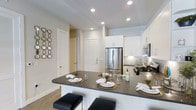 This single-floor residence is designed with a clean, contemporary aesthetic that prioritizes both style and functionality. The open-concept layout seamlessly connects the kitchen and living areas, creating an inviting and cohesive living environment. The kitchen is a standout feature, outfitted with sleek white cabinetry that extends to the ceiling, providing ample storage space. Stainless steel appliances, including a French door refrigerator, a gas range, and a built-in microwave, add a polished, modern touch. The dark countertops offer a striking contrast against the white cabinetry, and the peninsula-style breakfast bar comfortably accommodates seating for multiple guests, making it ideal for casual dining or entertaining. Recessed lighting throughout the kitchen ensures bright, even illumination. The light wood-toned flooring flows consistently across the space, contributing to a warm and airy ambiance. The neutral wall tones provide a versatile backdrop that complements a variety of décor styles. Decorative geometric wall accents in the adjoining living area add a touch of personality and visual interest. The space also includes interior doors with classic paneled detailing, lending a refined architectural quality to the overall design.