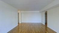 This single-floor space presents a straightforward and functional layout centered around an open main living area. The room is finished with warm-toned hardwood floors that extend throughout, adding a natural richness to the otherwise minimalist interior. The walls are painted in a clean, neutral white, creating a bright and airy atmosphere that allows the space to feel open and adaptable to various furnishing styles.

A structural beam detail is visible along the ceiling, subtly defining zones within the open area without disrupting the overall flow of the space. The ceiling is smooth and finished in white, contributing to the room's clean aesthetic. Toward the rear of the space, a doorway leads to an adjacent area, likely a hallway or secondary room, while built-in shelving is partially visible along the far right side, offering practical storage solutions.

The layout is unfurnished, making it easy to assess the full scale and versatility of the space. The combination of hardwood flooring, neutral finishes, and an open plan makes this a functional and adaptable space suitable for a variety of uses.