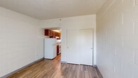Located at 11190 Mesquite Ave in Desert Hot Springs, CA, this efficient single-floor studio unit offers a practical and functional layout across four distinct areas: a living room, kitchen, bathroom, and closet. The living area features wood-look plank flooring, painted cinder block accent walls, and a textured ceiling, lending the space a clean, no-frills aesthetic. A wall-mounted HVAC unit and a window with vertical blinds provide climate control and natural light. A sliding double-door closet offers convenient in-room storage. The galley-style kitchen is accessible directly from the living area and is equipped with warm-toned wood cabinetry, a white refrigerator, and a gas range, all running along a single wall with a small window at the end for added brightness. The bathroom continues the wood-look flooring and includes a wall-mounted sink, toilet, overhead cabinetry, and accessibility grab bars. A mirror above the sink reflects the kitchen beyond, reinforcing the open and connected feel of the overall layout. The unit's compact design makes efficient use of every square foot, offering all essential amenities in a straightforward, easy-to-navigate floor plan.