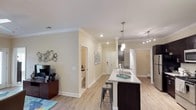 Located at 1415 E NC-54 Hwy in Durham, North Carolina, this single-floor residence offers a thoughtfully designed open-concept layout that seamlessly connects living and kitchen areas. The kitchen is a standout feature, showcasing a large island with granite countertops, a stainless steel sink, and stylish pendant lighting that adds a warm, contemporary feel. Dark cabinetry provides a striking contrast against the light stone counters, while recessed lighting and track lighting ensure the space is well-illuminated throughout. Light hardwood flooring runs continuously across the main living areas, enhancing the sense of openness and creating a cohesive, inviting atmosphere. Crown molding adds an elegant architectural detail along the ceilings, contributing to the polished look of the interior. Neutral, warm-toned walls complement the finishes and provide a versatile backdrop throughout the home. The hallway leads to additional rooms, including what appears to be a bathroom, offering a functional and well-organized floor plan. The living area accommodates comfortable furnishings, and decorative elements like wall art contribute to the overall welcoming character of this well-appointed single-level home.