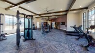 This single-floor fitness center is thoughtfully designed to offer a comprehensive workout experience. The space is anchored by patterned, multi-tone carpet flooring throughout, complemented by recessed lighting and ceiling fans that maintain a comfortable environment during exercise. Exposed dark wood beams accent the vaulted ceiling areas, adding an architectural touch to the athletic setting.

The cardio zone is positioned along a wall of expansive floor-to-ceiling grid-style windows, providing abundant natural light and unobstructed views of an outdoor pool and surrounding community. This section houses treadmills, elliptical machines, stationary bikes, and a rowing machine, allowing members to exercise while taking in the scenery. A mounted flat-screen television accompanies this area.

A dedicated strength training section features multiple cable and resistance machines, adjustable benches, a dumbbell rack, and weight plates. A standout feature is the Freedom Climber — a motorized climbing wall — which serves as a unique addition to the gym's functional fitness offerings. A functional training rig with suspension straps, battle ropes, and medicine balls rounds out the versatile equipment selection.

A decorative topographic map mural referencing Western N.C. Trailheads adds a regional, outdoor-inspired aesthetic to one accent wall, tying the space's design together with a sense of adventure and place.