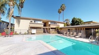 This single-story property showcases a well-appointed outdoor communal area designed for relaxation and social gathering. The centerpiece is a generously sized swimming pool with clean, turquoise water, framed by a concrete pool deck lined with lounge chairs — ideal for sunbathing and leisure. A wrought-iron security fence encloses the pool area, providing both safety and a defined boundary for the space.

Adjacent to the pool, a shaded patio area features a canopy gazebo covering an outdoor dining table with red chairs, offering a comfortable spot for al fresco meals or casual gatherings. A barbecue grill is also available nearby, adding to the space's entertainment appeal. The patio is finished with a concrete surface and bordered by decorative iron railings.

The surrounding landscape is characterized by tall, mature palm trees and lush tropical greenery that contribute to a warm, Southern California-style ambiance. The multi-building exterior features a neutral stucco finish with covered walkways and stairwell access. Under consistently clear, sunny skies, the outdoor environment feels open, bright, and inviting throughout the day.