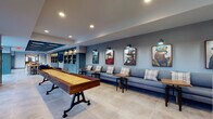 This single-floor amenity space is thoughtfully designed to blend social, recreational, and functional areas into one cohesive environment. The layout flows seamlessly from an entry corridor—featuring teal-painted built-in shelving and accent doors—into a spacious lounge anchored by a full-length shuffleboard table with a rich wood surface and industrial iron base. Along one wall, a continuous gray sectional sofa is paired with plaid throw pillows and side tables, while a series of whimsical anthropomorphic animal portraits adds personality and visual interest. Recessed lighting and wall-mounted sconces keep the atmosphere warm throughout.

Beyond the lounge, the space opens to a well-appointed kitchen area featuring a striking waterfall-edge marble island with bar-height seating, pendant lighting, subway tile backsplash, and stainless steel appliances. Clerestory windows allow natural light to filter in from multiple directions, and a large floor-to-ceiling window at one end frames an outdoor view of mature trees. Porcelain tile flooring unifies the main areas, while a private meeting nook with warm hardwood flooring, a marble-top table, and patterned upholstered chairs offers a quieter retreat with its own natural light.