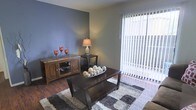 This single-floor space presents a well-appointed living area that balances comfort and modern style. The room is anchored by rich hardwood flooring that adds warmth and character throughout. A striking deep-gray accent wall serves as a bold focal point, creating a sense of depth and contrast against the lighter tones of the surrounding walls. Large sliding glass doors dressed with vertical blinds allow an abundance of natural light to pour into the space, enhancing its open and airy feel while offering a direct connection to the outdoors. The living area is thoughtfully laid out, with ample room for a central coffee table set atop a plush, multi-toned area rug that adds texture and visual interest underfoot. A substantial wooden media console lines one wall, providing both storage and display space, complemented by warm decorative accents. The overall ambiance is inviting and contemporary, with a cohesive blend of dark wood furnishings and neutral tones that create a grounded, livable aesthetic. The space is well-suited for both everyday living and casual entertaining, with a layout that feels both functional and comfortable.