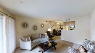 This single-story residence is designed with an open-concept layout that seamlessly connects the living room, dining area, and kitchen into one cohesive living space. The living room is carpeted and anchored by a curved, light-toned sofa paired with a dark wood coffee table, creating a comfortable and welcoming atmosphere. Warm beige walls are accented with decorative sunburst mirrors, adding a stylish focal point to the room. A textured popcorn ceiling runs throughout, contributing to the space's cozy character. Natural light fills the area, enhancing the warm neutral color palette.

The dining area sits just beyond the living room, accommodating a dark wood dining table and chairs beneath a pendant light fixture, ideal for everyday meals or casual gatherings. It transitions directly into the kitchen, which features medium-toned wood cabinetry, a raised breakfast bar, and standard appliances. The kitchen is efficiently laid out, offering ample counter and storage space. Overall, the space presents a functional and inviting interior with a consistent warm tone and practical open-plan design throughout.