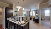 The Cortona is a thoughtfully designed 2-bedroom, 2-bathroom residence that combines modern finishes with a functional, open-concept layout. The kitchen serves as the heart of the home, featuring dark espresso cabinetry, granite countertops, a tile backsplash, black appliances, and a breakfast bar illuminated by stylish pendant lighting. Rich hardwood-style flooring flows through the kitchen and extends into both bathrooms, while plush carpet adds warmth to the living areas and bedrooms. Crown molding accents the main living spaces, lending an elevated sense of sophistication. The open dining area accommodates a full dining set and connects seamlessly to the living room, which enjoys natural light from multiple windows dressed with elegant drapery. Ceiling fans are present in both the living room and bedrooms for added comfort. Each bedroom offers its own walk-in closet, providing generous storage. Both bathrooms are appointed with dark cabinetry, granite vanity tops, and walk-in showers. The primary bathroom features an especially spacious layout with a large walk-in closet accessible directly from the bath. Throughout the residence, the warm neutral palette and consistent design details create a cohesive, inviting ambiance ideal for comfortable everyday living.