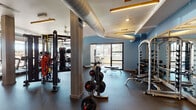 Located at 2337 S Blackhawk St in Aurora, Colorado, this well-appointed fitness center offers a comprehensive workout environment designed to accommodate a wide range of exercise needs. The facility is organized into distinct functional zones, each thoughtfully equipped and finished.

The main workout floor features durable rubber flooring and is outfitted with an extensive selection of free weights, including barbells, weight plates, and a multi-function cable and suspension training station complete with a heavy punching bag. A dedicated dumbbell rack and adjustable benches provide ample space for strength training. Ceiling fans and exposed ventilation ductwork contribute to an industrial-modern aesthetic, while warm linear pendant lighting adds a polished touch against the blue accent walls.

A separate cardio zone lines the perimeter of the space with a row of treadmills, ellipticals, and other cardio machines positioned to face floor-to-ceiling windows, offering views of the surrounding outdoors and filling the space with natural light.

A dedicated studio room features hardwood flooring, full-length mirrors, a ballet barre, and a flat-screen TV, making it ideal for group classes, yoga, or individual training sessions. Medicine balls, plyo boxes, and balance trainers are neatly organized within the studio. A small service counter and storage cubbies near the main floor provide convenient amenity support for facility users.