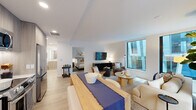 This thoughtfully designed single-floor residence offers a seamless blend of modern aesthetics and functional living. The entryway welcomes guests with light wood flooring, a circular mirror, and a sleek console, setting a warm yet contemporary tone throughout. The open-concept living area is anchored by large floor-to-ceiling windows that invite abundant natural light and offer views of the surrounding cityscape. The living space is furnished with a curated mix of seating, a wall-mounted television atop a black media console, and a dining area with a natural wood table accented by a navy runner. The kitchen is fitted with light wood cabinetry extending to the ceiling, white countertops with under-cabinet lighting, and a full suite of stainless steel appliances including a French door refrigerator, range, microwave, and dishwasher. The bedroom is a serene retreat featuring a large window with city views, a built-in closet with sliding doors, and warm accent lighting. The bathroom offers a clean, minimal design with a glass-enclosed walk-in shower, wood-toned vanity cabinetry, and tile flooring. Light wood floors flow consistently throughout, unifying the space with a cohesive, contemporary character.