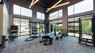 Located at 3700 National Dr in Raleigh, NC, this two-story fitness facility delivers a comprehensive workout environment defined by a bold industrial aesthetic. The design pairs dark exposed ceilings and structural columns with geometric linear lighting fixtures, creating a sleek, high-energy atmosphere throughout.

The main level's centerpiece is a soaring double-height functional training area, where floor-to-ceiling glass walls flood the space with natural light and offer open views of the surrounding greenery. This zone is equipped with a multi-functional rig, kettlebells, battle ropes, a punching bag, medicine balls, and a tire sled, accommodating a wide range of strength and conditioning workouts. A metal staircase with industrial railings connects the two floors, reinforcing the facility's architectural character.

Additional training areas house rows of treadmills, ellipticals, stationary bikes, and rowing machines, with large mirrors amplifying the sense of space. A dedicated studio room features light hardwood flooring, full-length mirrored walls, a ceiling fan, built-in storage, and a large display screen—ideal for group classes or personal training sessions. Throughout, textured carpet tile flooring and curated artwork contribute to a polished yet functional environment.