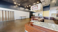 Located at 13676 Noel Rd in Dallas, TX, Suite A22 is a single-floor commercial space offering a thoughtfully arranged layout across a generous footprint. The front reception area makes a strong first impression, featuring polished concrete floors, soaring ceilings with dark upper walls, and a mix of pendant and track lighting that creates a dynamic, upscale atmosphere. A bold, colorful mural near the storefront windows adds a vibrant artistic focal point, while large windows invite abundant natural light and street views.

At the heart of the space is a prominent bar-style counter with rich wood cabinetry and granite-style countertops, complemented by a striking beverage-themed graphic wall mural that reinforces a hospitality-oriented identity. A textured decorative wall panel and a wall-mounted shelf further enhance the front area's character.

Beyond the main area, a series of hallways connect to back-of-house zones equipped with utility infrastructure, including a commercial sink, floor drains, exposed plumbing, and storage partitions with metal cabinetry. These functional areas are finished with durable tile flooring suited for operational demands. The overall layout supports a variety of commercial uses, particularly those in the food, beverage, or personal services industries.