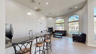 Located at 5150 N 99th Ave in Phoenix, Arizona, this single-story community clubhouse offers a thoughtfully designed layout spread across a generously sized floor plan. The space is defined by its striking architectural character, including soaring ceilings, arched windows, and light hardwood flooring that flows throughout.

The main lounge area is a welcoming centerpiece, featuring tall arched windows that flood the room with natural light and frame views of the outdoor pool and surrounding landscaping. Dark leather sofas surround a wood coffee table, and a stone-accented fireplace adds warmth and visual interest. Pendant lighting illuminates a granite-topped wet bar with barstool seating, ideal for casual gatherings.

An adjacent dining area provides multiple high-top tables and chairs beneath arched windows and a wall-mounted TV, creating a versatile space for socializing or community events. Large glass doors open directly to the outdoor patio, enhancing the connection to the exterior.

The second main area is a dedicated home theater room, finished in cool gray tones with ambient LED base lighting. Tiered rows of black leather reclining seats with cup holders face a large wall-mounted screen, while movie posters and wall-mounted speakers complete the cinematic atmosphere. The grand entry foyer with its glass-paneled French doors ties the entire space together with elegant simplicity.