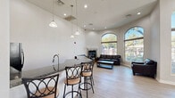 Located at 5150 N 99th Ave in Phoenix, Arizona, this single-story community clubhouse offers a thoughtfully designed layout spread across a generously sized floor plan. The space is defined by its striking architectural character, including soaring ceilings, arched windows, and light hardwood flooring that flows throughout.

The main lounge area is a welcoming centerpiece, featuring tall arched windows that flood the room with natural light and frame views of the outdoor pool and surrounding landscaping. Dark leather sofas surround a wood coffee table, and a stone-accented fireplace adds warmth and visual interest. Pendant lighting illuminates a granite-topped wet bar with barstool seating, ideal for casual gatherings.

An adjacent dining area provides multiple high-top tables and chairs beneath arched windows and a wall-mounted TV, creating a versatile space for socializing or community events. Large glass doors open directly to the outdoor patio, enhancing the connection to the exterior.

The second main area is a dedicated home theater room, finished in cool gray tones with ambient LED base lighting. Tiered rows of black leather reclining seats with cup holders face a large wall-mounted screen, while movie posters and wall-mounted speakers complete the cinematic atmosphere. The grand entry foyer with its glass-paneled French doors ties the entire space together with elegant simplicity.