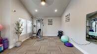 This single-story fitness center offers a spacious, open layout designed to support a well-rounded workout routine. The room's most distinctive architectural feature is its vaulted ceiling, which rises dramatically to create an airy, open atmosphere enhanced by recessed lighting and a ceiling fan that promotes airflow during exercise. Neutral-toned carpet tiles cover the floor throughout, providing a clean, functional surface across the entire space.

The facility is divided into two primary functional zones. The cardio area is equipped with a treadmill, elliptical, and stationary spin bike, positioned near large windows and glass-panel exterior doors that allow abundant natural light and views of the surrounding outdoor area. A second exit door provides convenient access to the exterior.

The strength and flexibility zone features a multi-function cable machine with an adjustable bench, a rack of dumbbells in various weights, stability balls, a BOSU balance trainer, resistance bands, and a yoga mat storage rack. A potted tree adds a natural accent to the space. Motivational wall decals — including "Don't Stop When You're Tired, Stop When You're Done" and "Pain Is Temporary, Quitting Lasts Forever" — reinforce an energizing atmosphere. A water station is also present, adding everyday convenience for users.