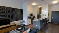 This single-floor space showcases a thoughtfully designed open-concept layout that seamlessly connects the living area and kitchen. The kitchen is outfitted with stainless steel appliances, including a range, microwave, and refrigerator, complemented by crisp white cabinetry and light stone countertops. A bold gray peninsula with seating extends into the main living area, topped with pendant lighting that adds a modern, polished touch. The floors throughout feature wood-style planking in a warm tone, providing a cohesive visual foundation. The living area is anchored by a patterned accent wall featuring a striking black-and-white geometric design, adding a focal point to the space. Recessed and flush-mount ceiling fixtures provide ample ambient lighting throughout. The layout makes efficient use of the available space, with a small side table and media console tucked neatly along the walls. Overall, the unit presents a clean, contemporary aesthetic with a neutral color palette punctuated by bold design choices, making the space feel both functional and visually engaging.